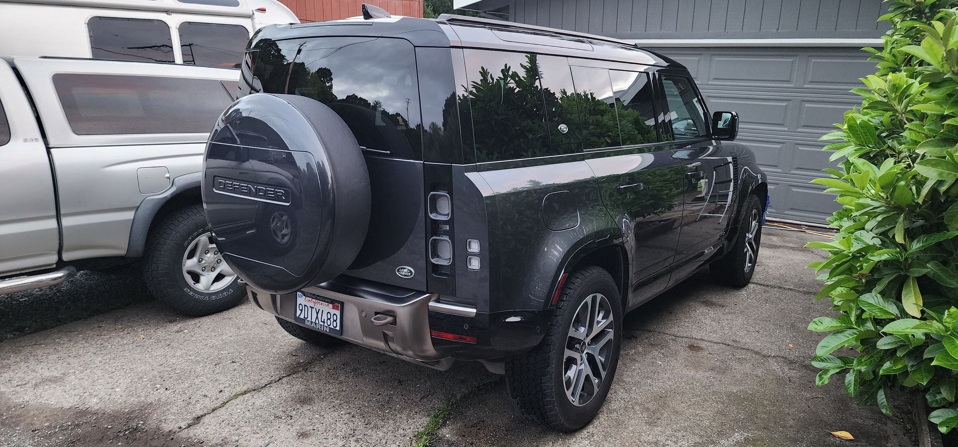 Black SUV parked in a driveway beside a silver vehicle and green bushes, with a spare tire cover on the back