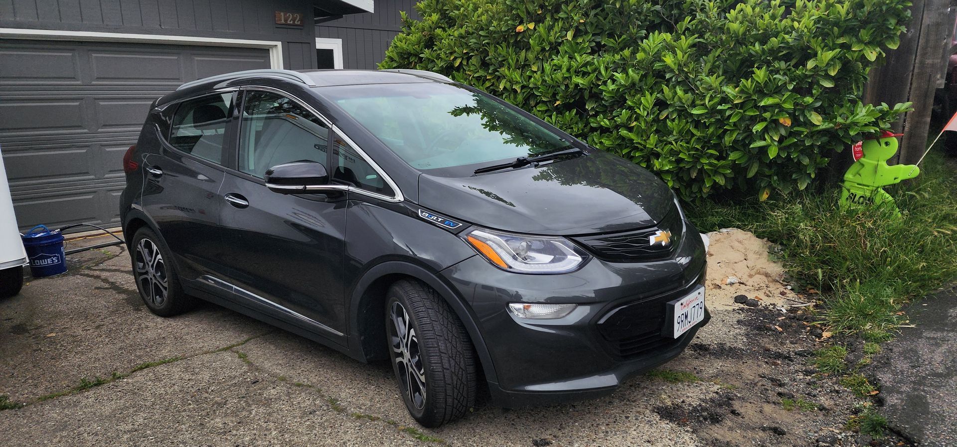 Dark gray hatchback car parked on a gravel driveway beside a garage and green bushes.