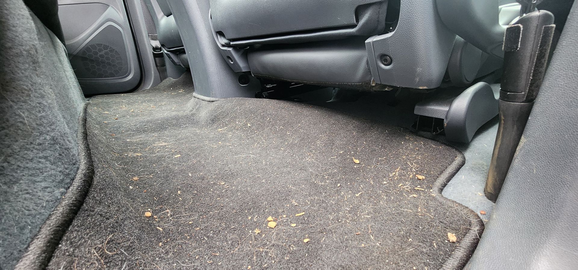 Car interior with gray carpeted floor covered in crumbs and debris, between the front seats and back seat