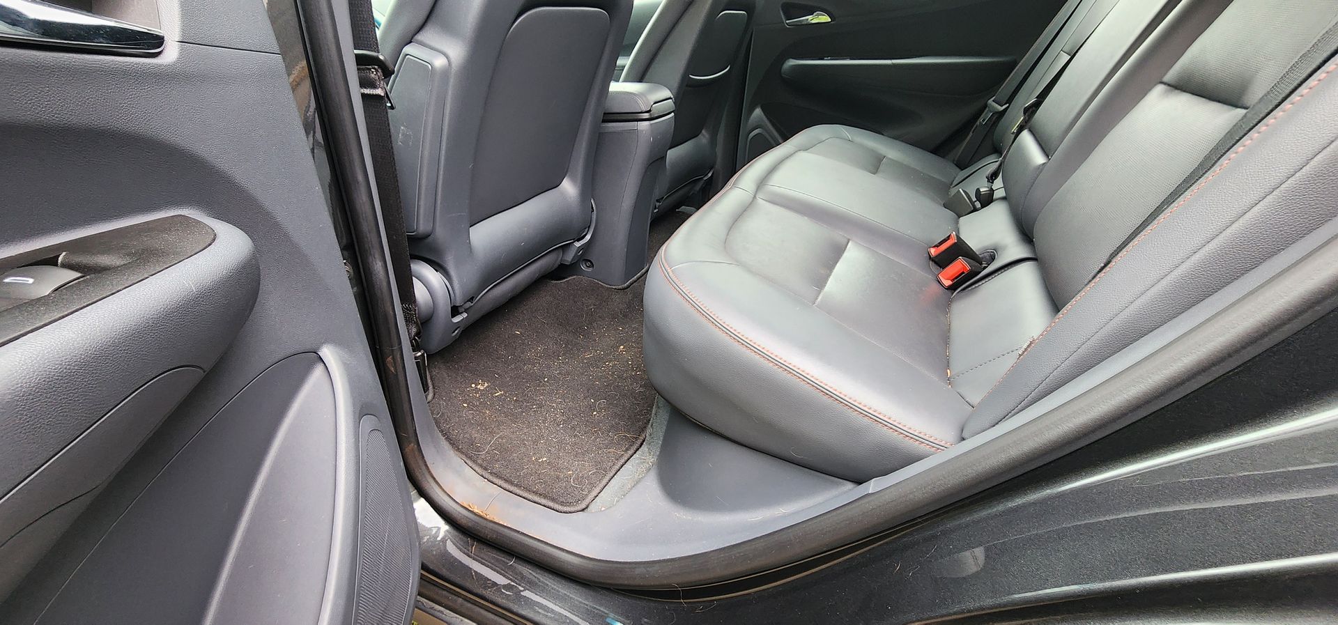Car interior with open rear door and gray fabric back seats, showing seat belts and floor mat.