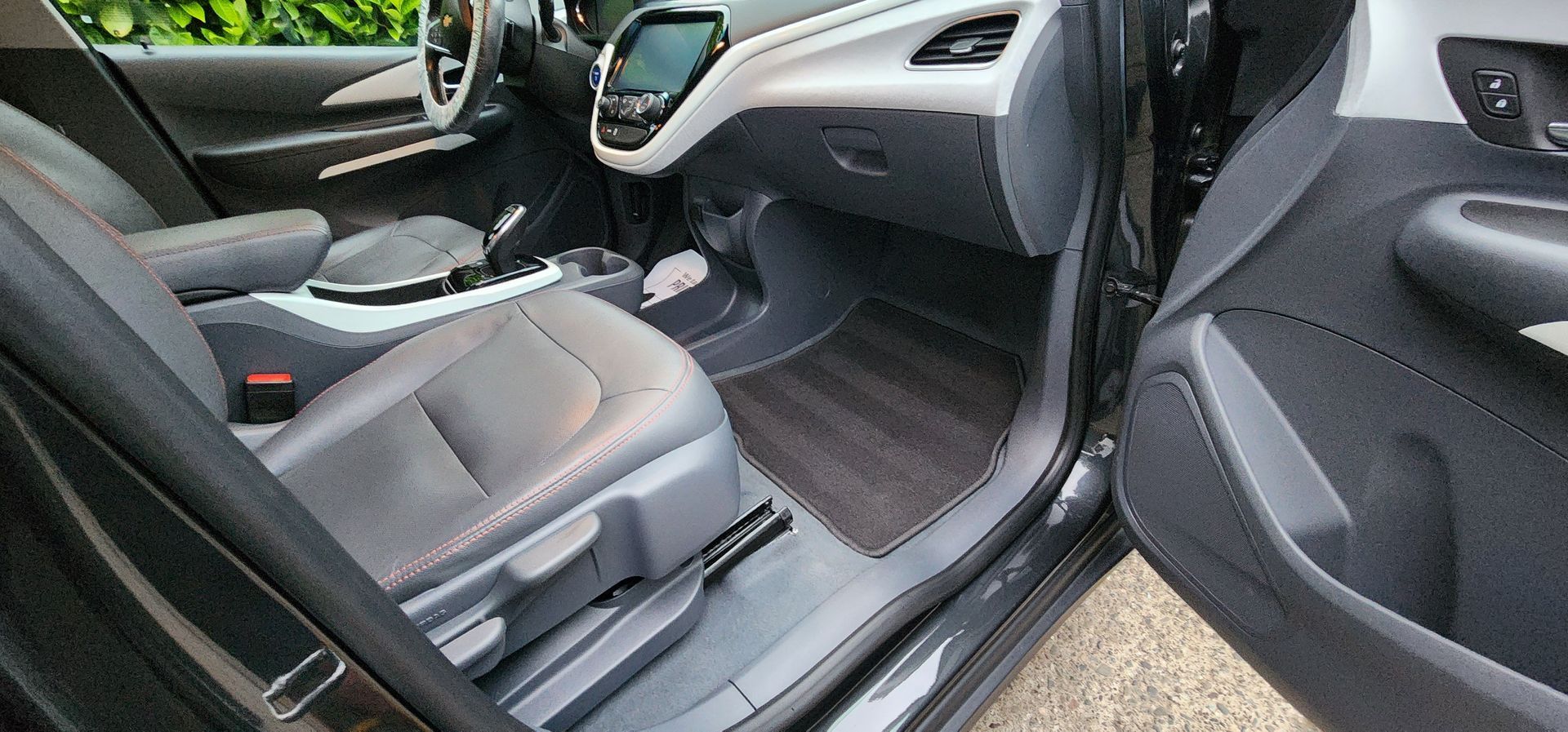 Car interior with black front seats, dashboard, and open passenger door on a driveway