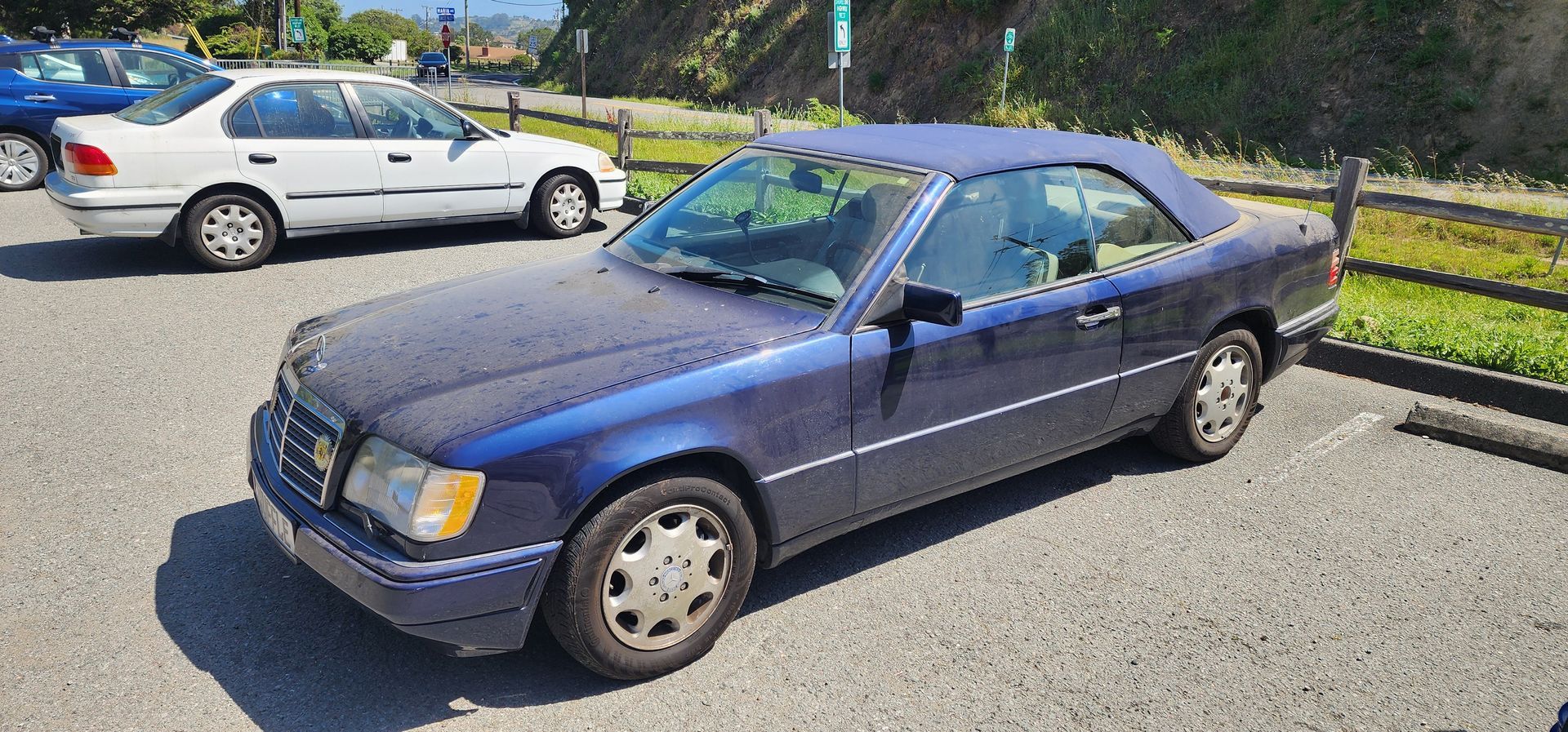 Blue two-door car parked on a gravel lot in bright sunlight, with a white car in the background.