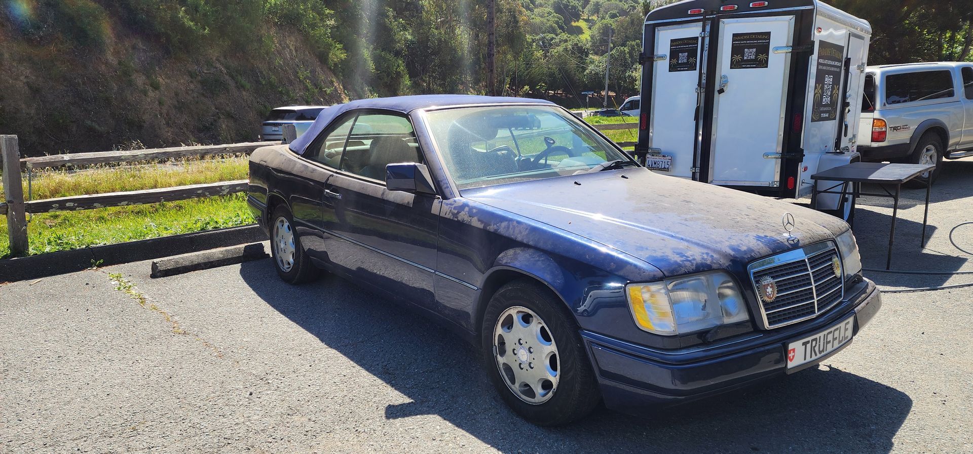 Dark blue sedan parked outdoors beside a trailer on a sunny day