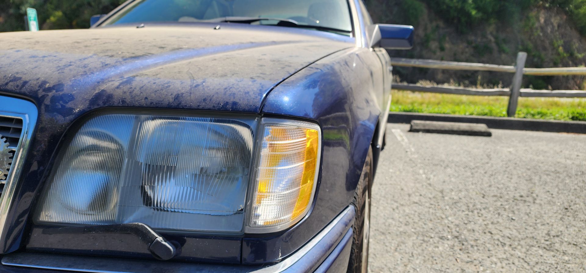 Close-up of a blue car’s front headlight and hood, parked by a road with a wooden fence in the background.