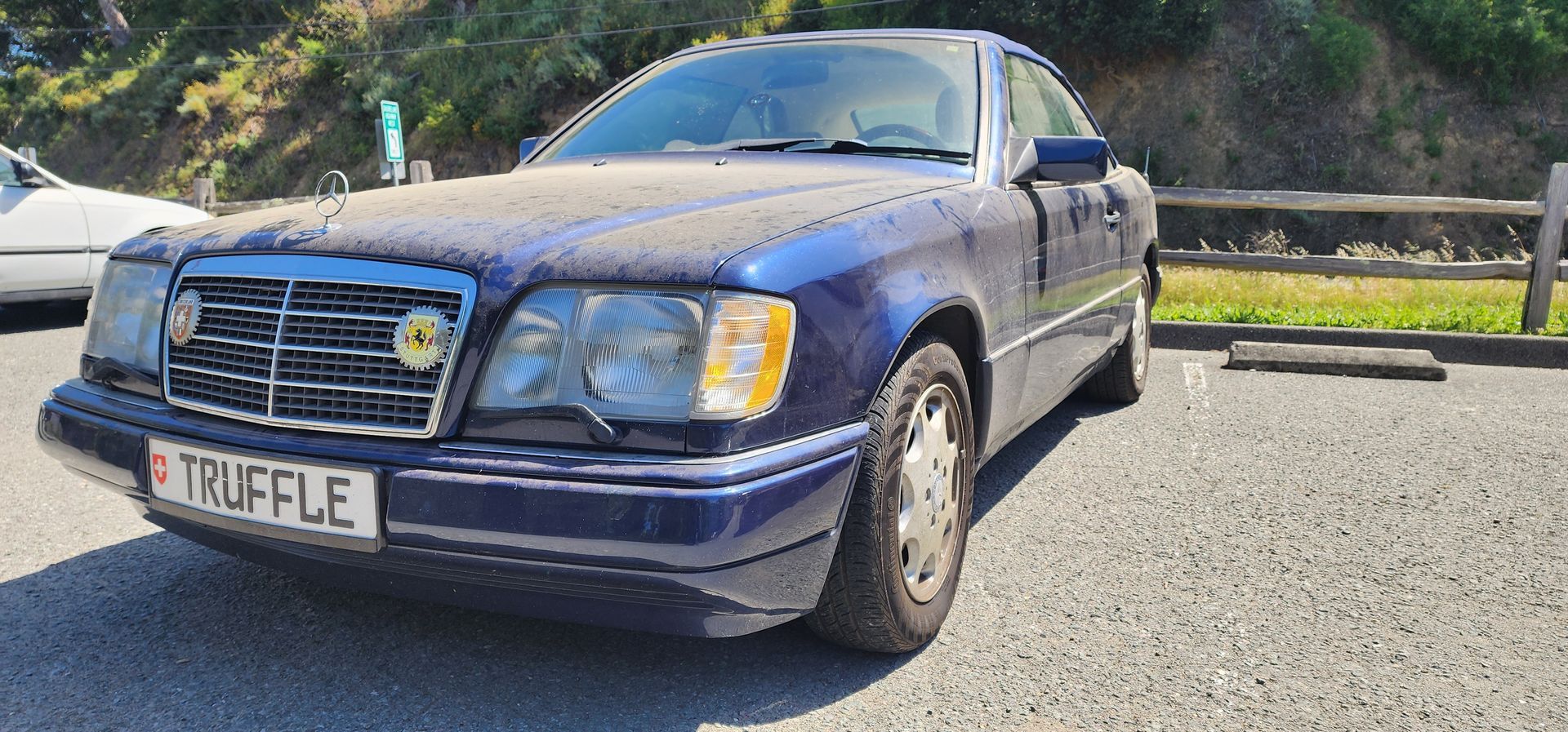 Blue sedan parked on a roadside in bright sunlight, front-left view with hills in the background