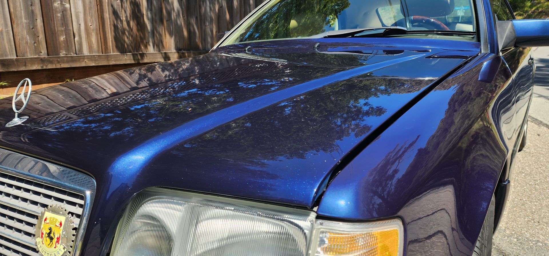 Blue car parked outdoors in front of a wooden fence, sunlight reflecting on the hood