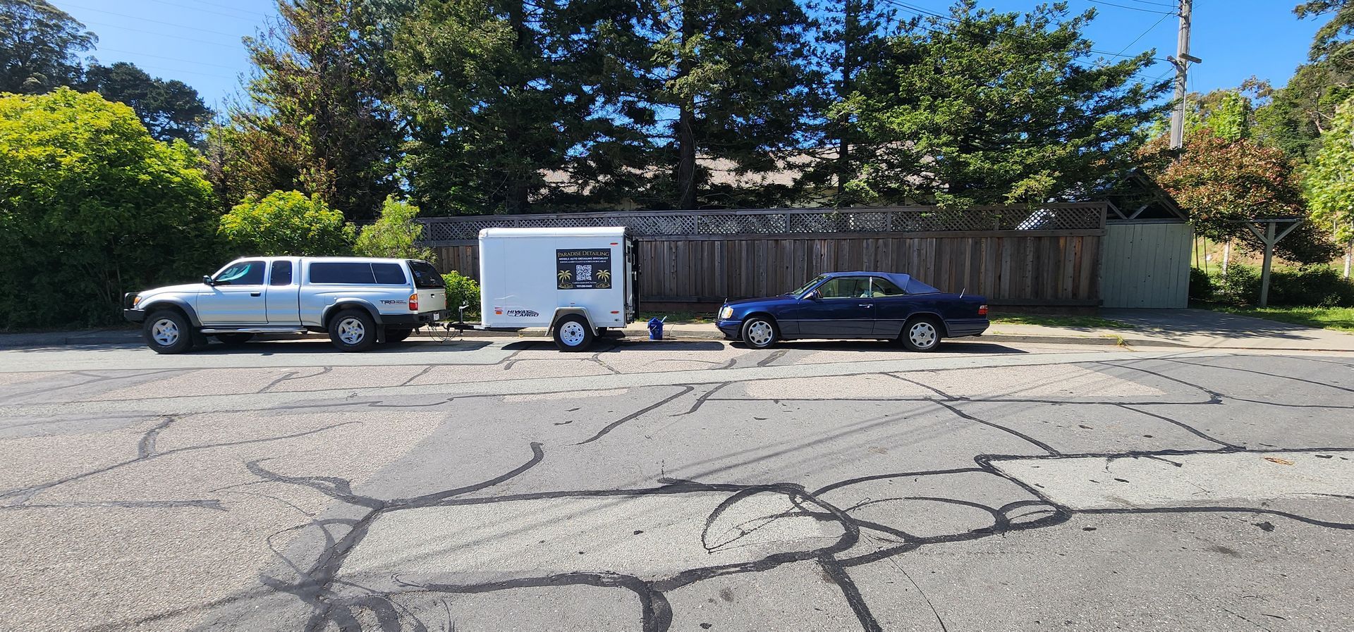Parked white SUV and black sedan beside a small white trailer in a cracked parking lot under trees.