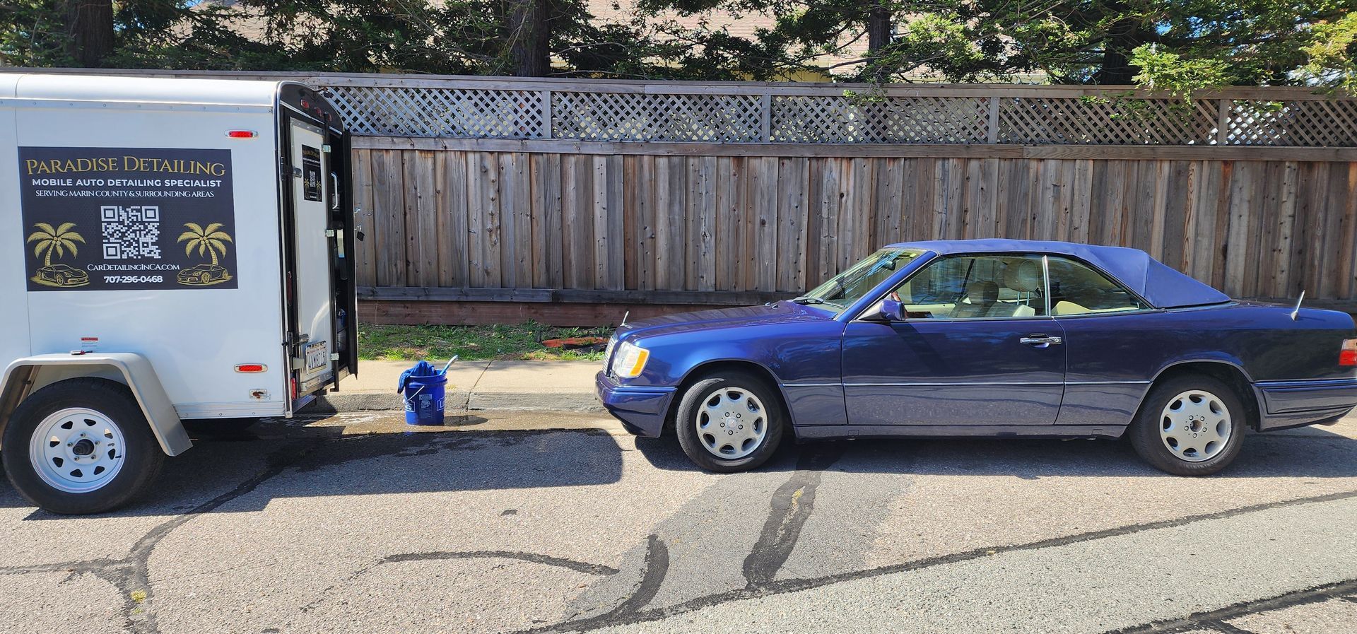 Blue coupe parked behind a white trailer on a street, with a wooden fence in the background.