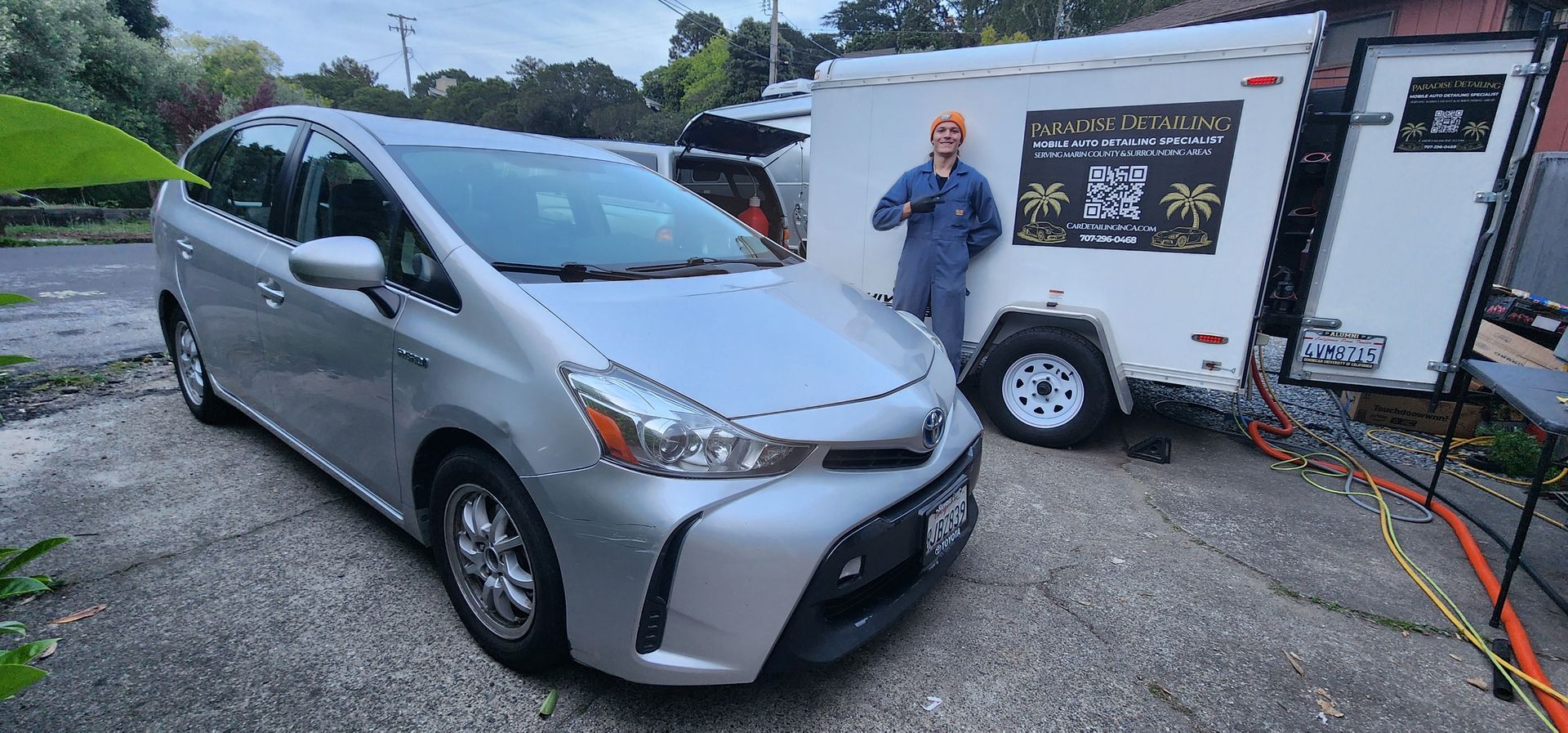 Silver minivan charging at a roadside station beside a trailer with a man standing nearby