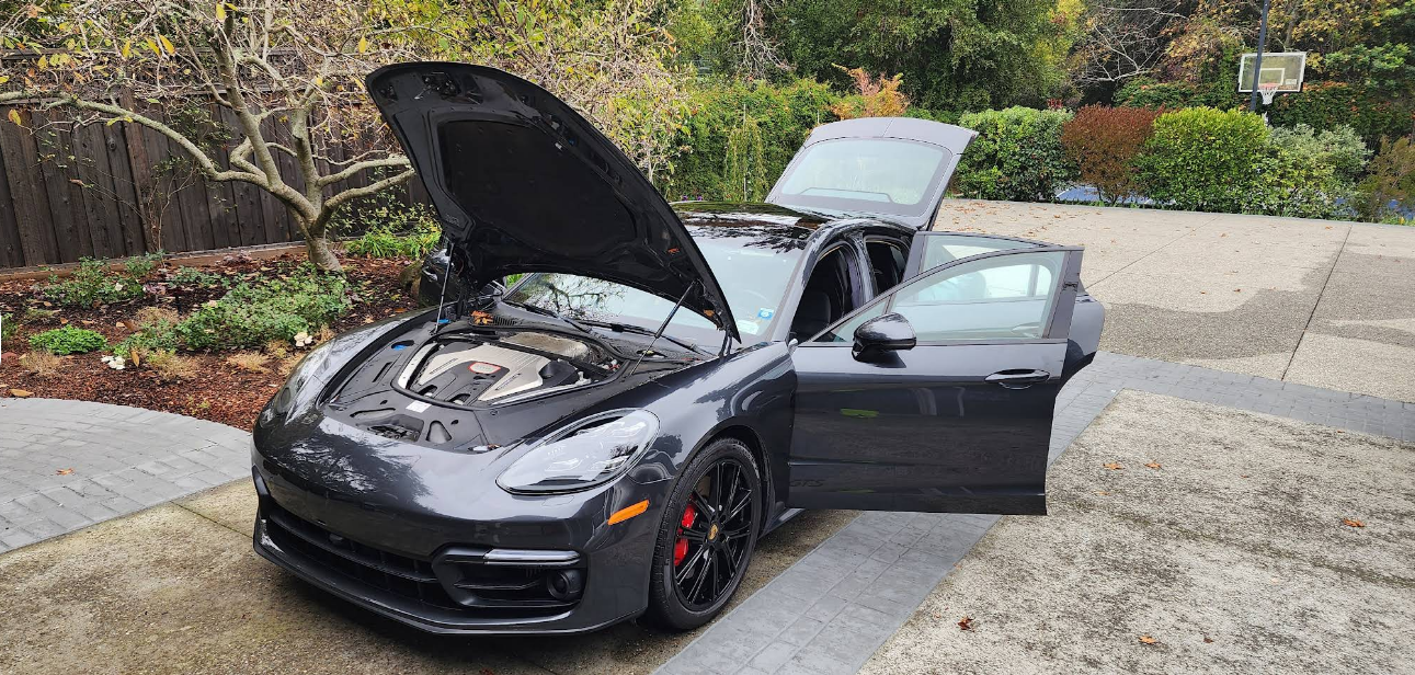 Black sports car with hood and trunk open, parked on a driveway beside greenery