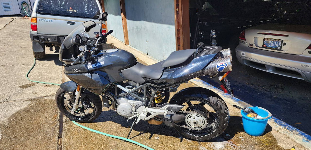 Black motorcycle parked in a driveway, being washed with a blue bucket beside it.