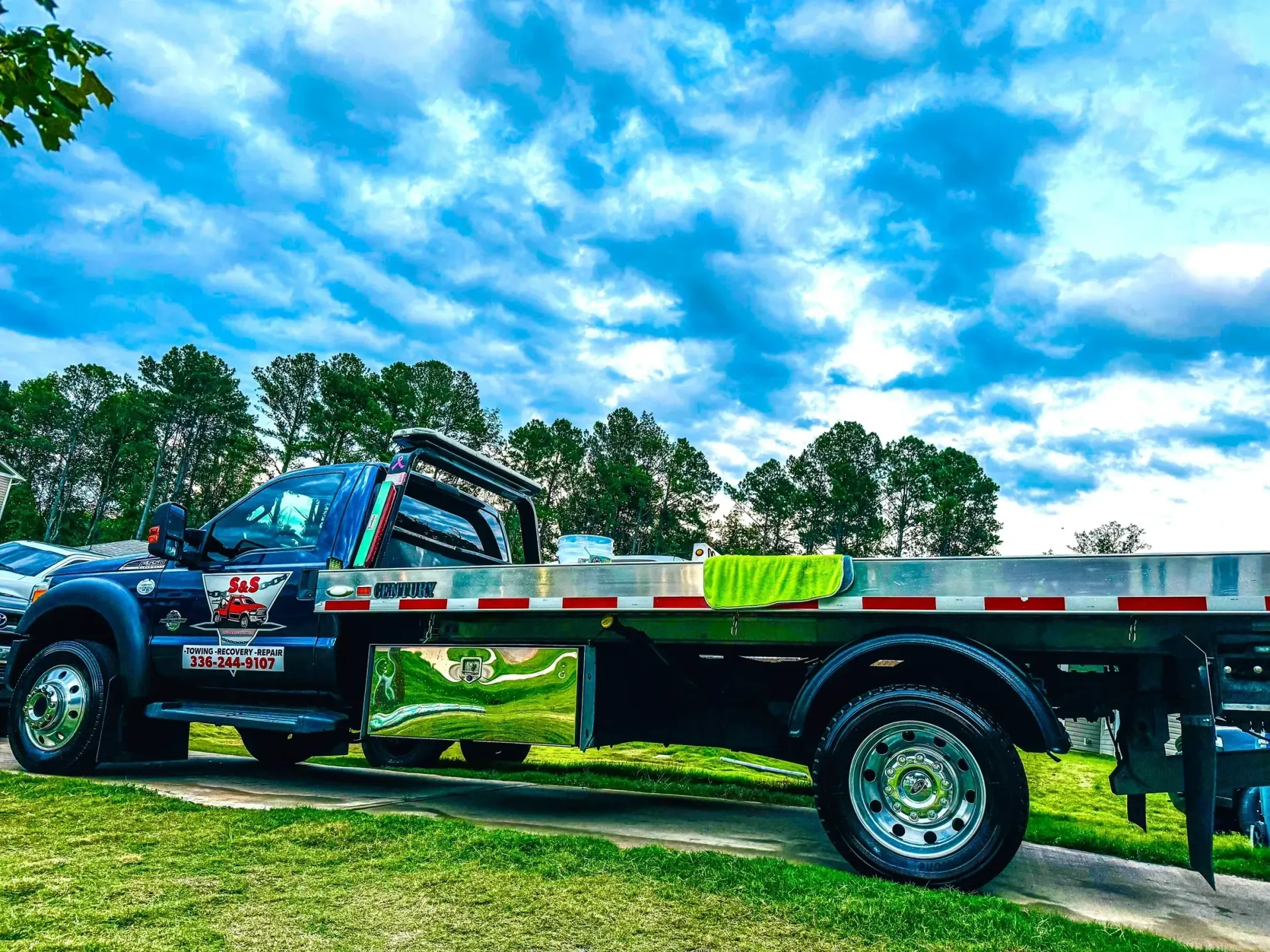 Flatbed tow truck parked on grass under a bright blue sky with clouds