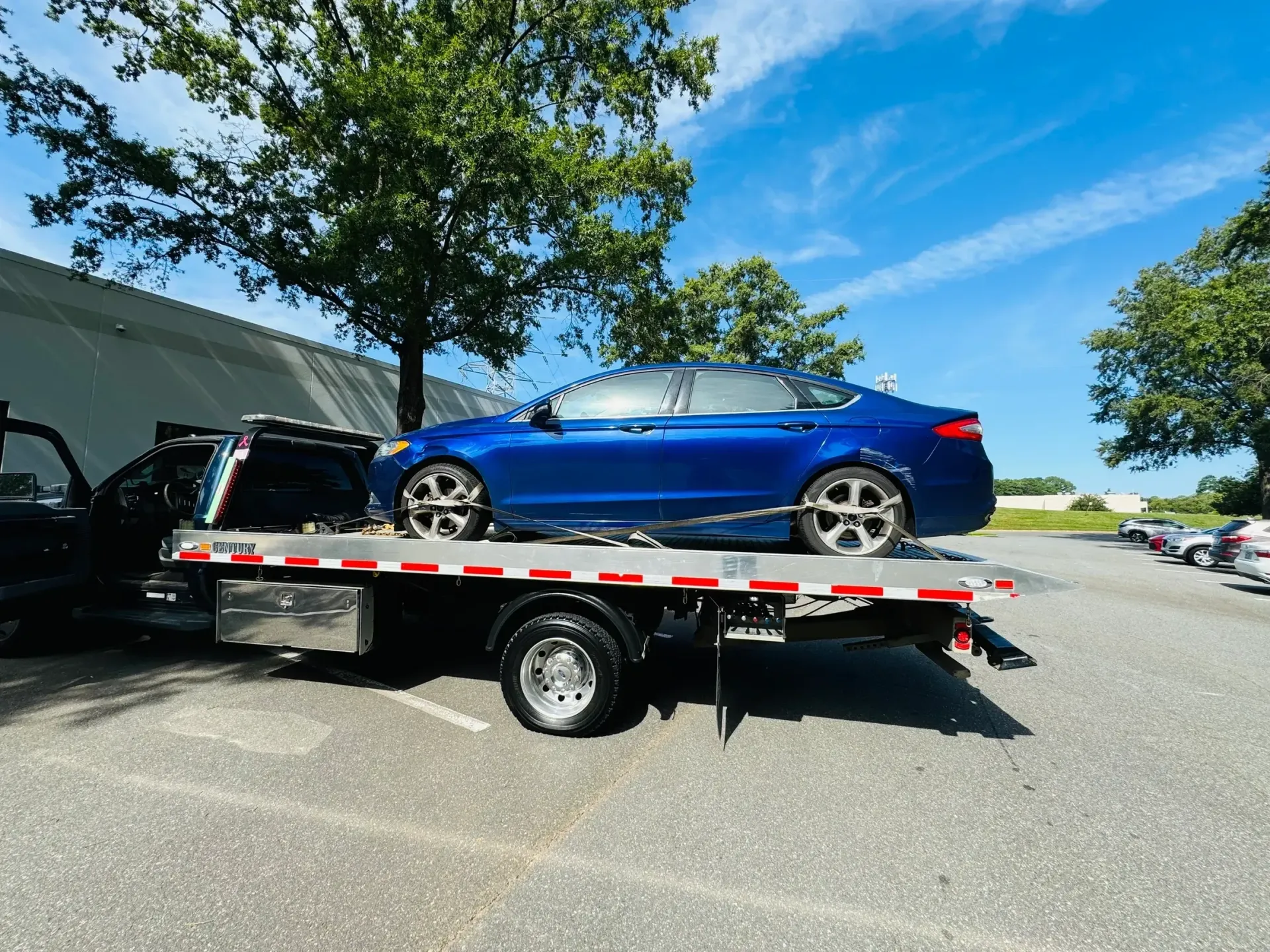 Blue car strapped on a flatbed tow truck in a parking lot under a sunny sky.