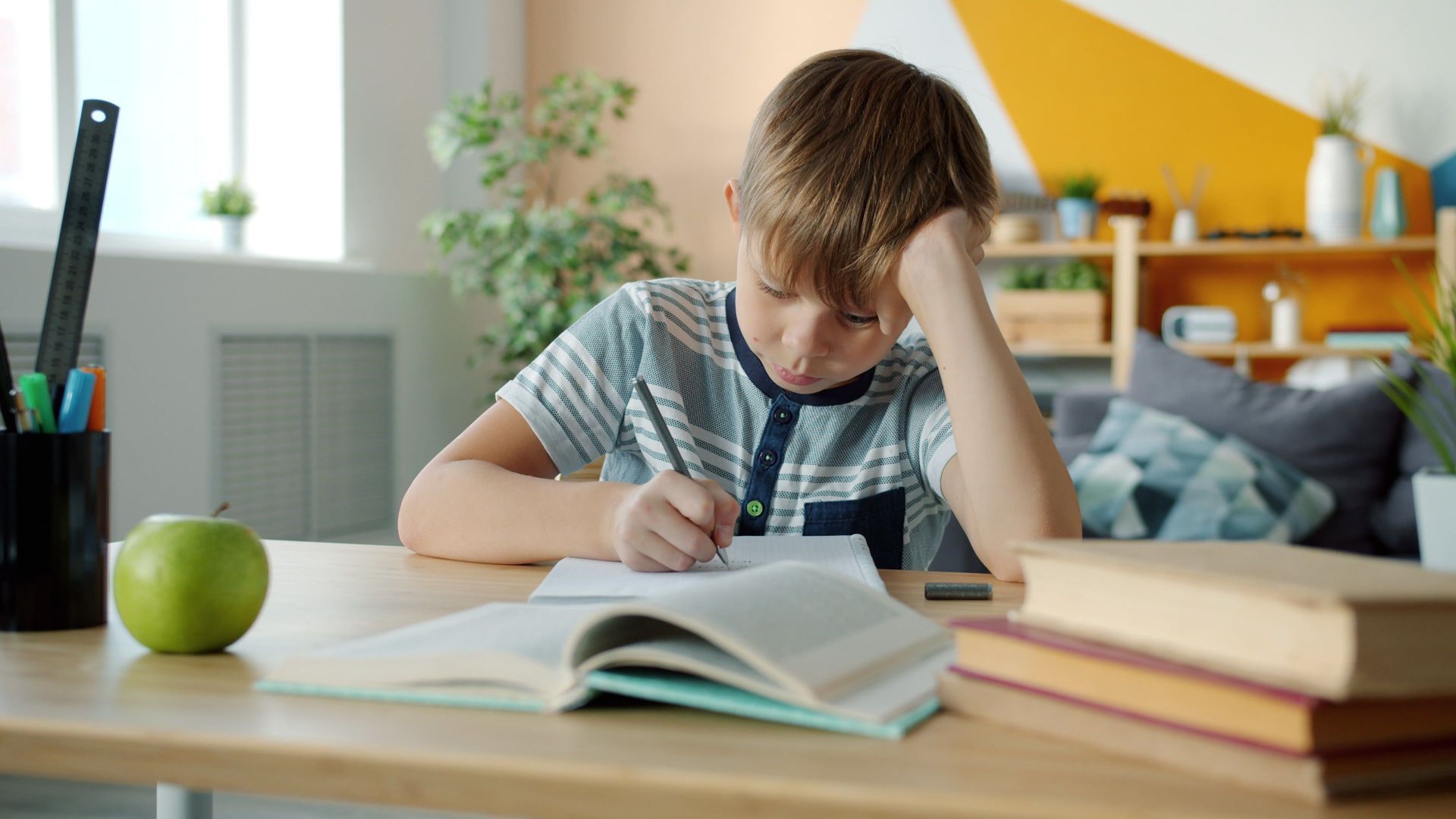 Child studying at a desk, writing in an open notebook with books, a green apple, and a laptop nearby.
