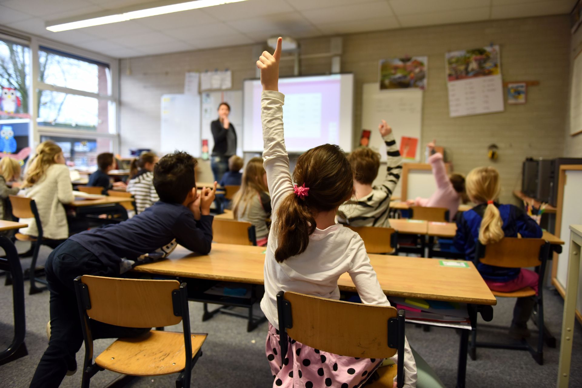Classroom of students raising hands while teacher stands at the front near a whiteboard