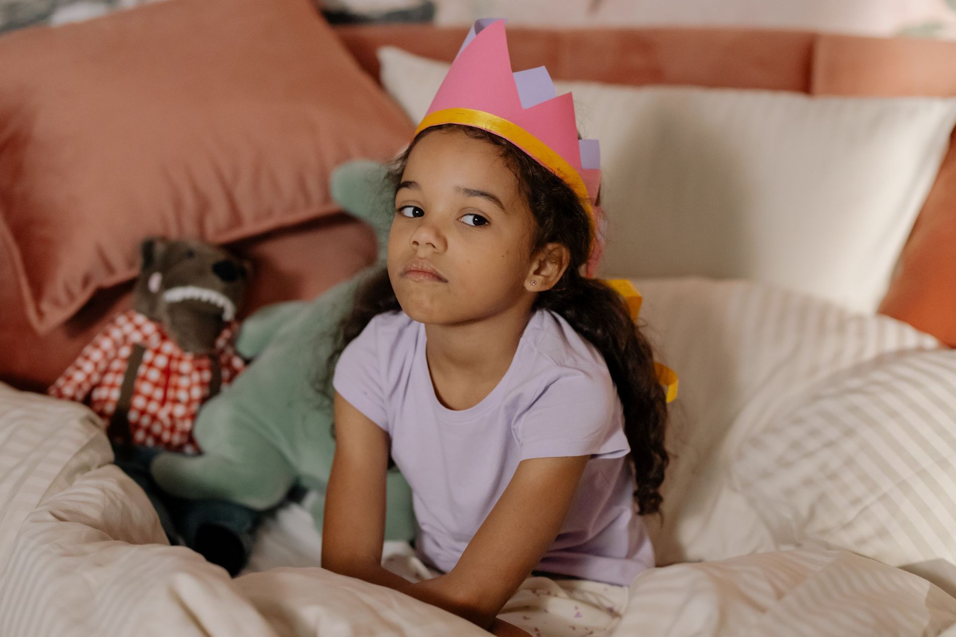 Child in a pink paper crown sits on a bed, wearing a lavender shirt and looking at the camera.