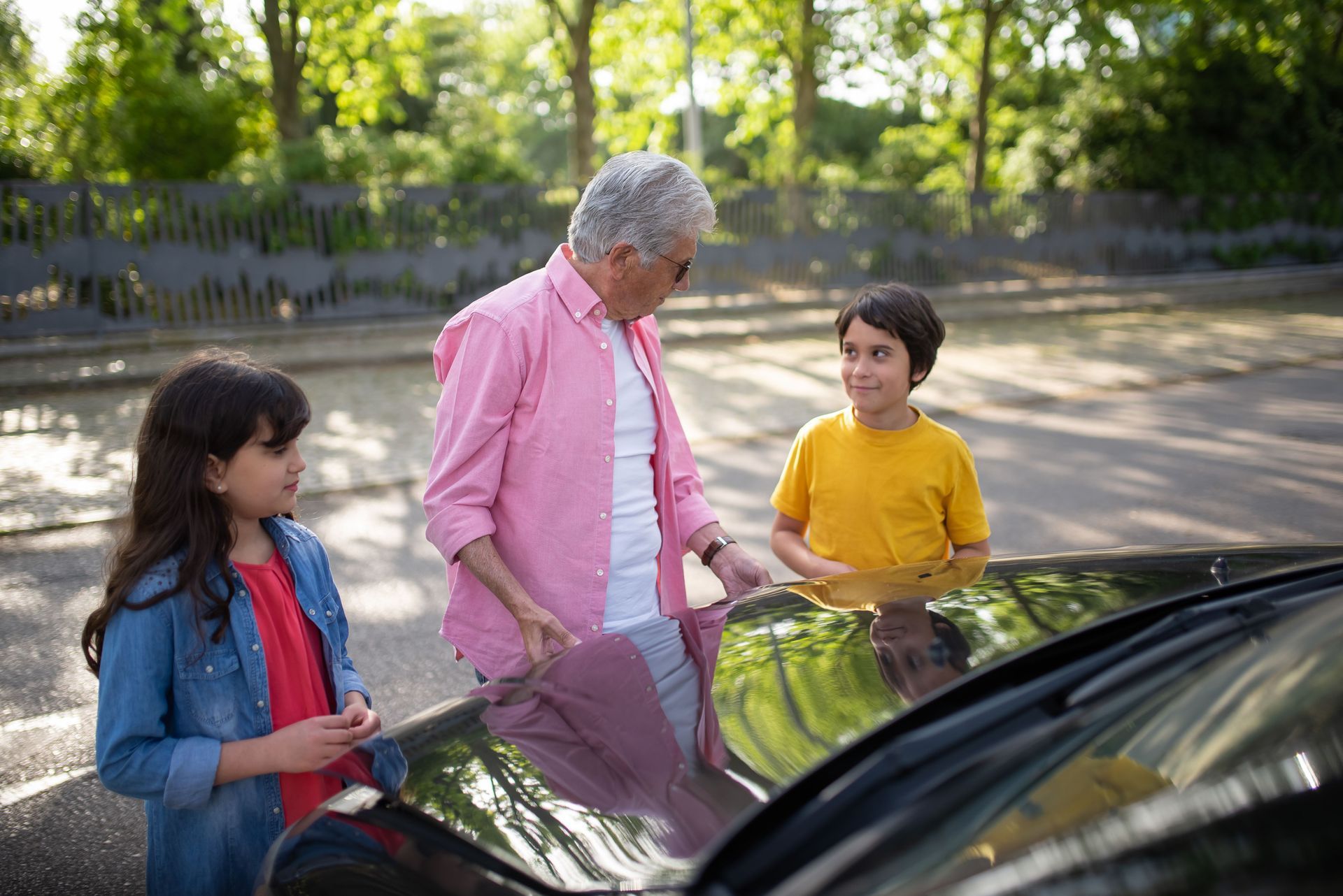 Three people stand by an open car door outdoors, looking inside and talking.