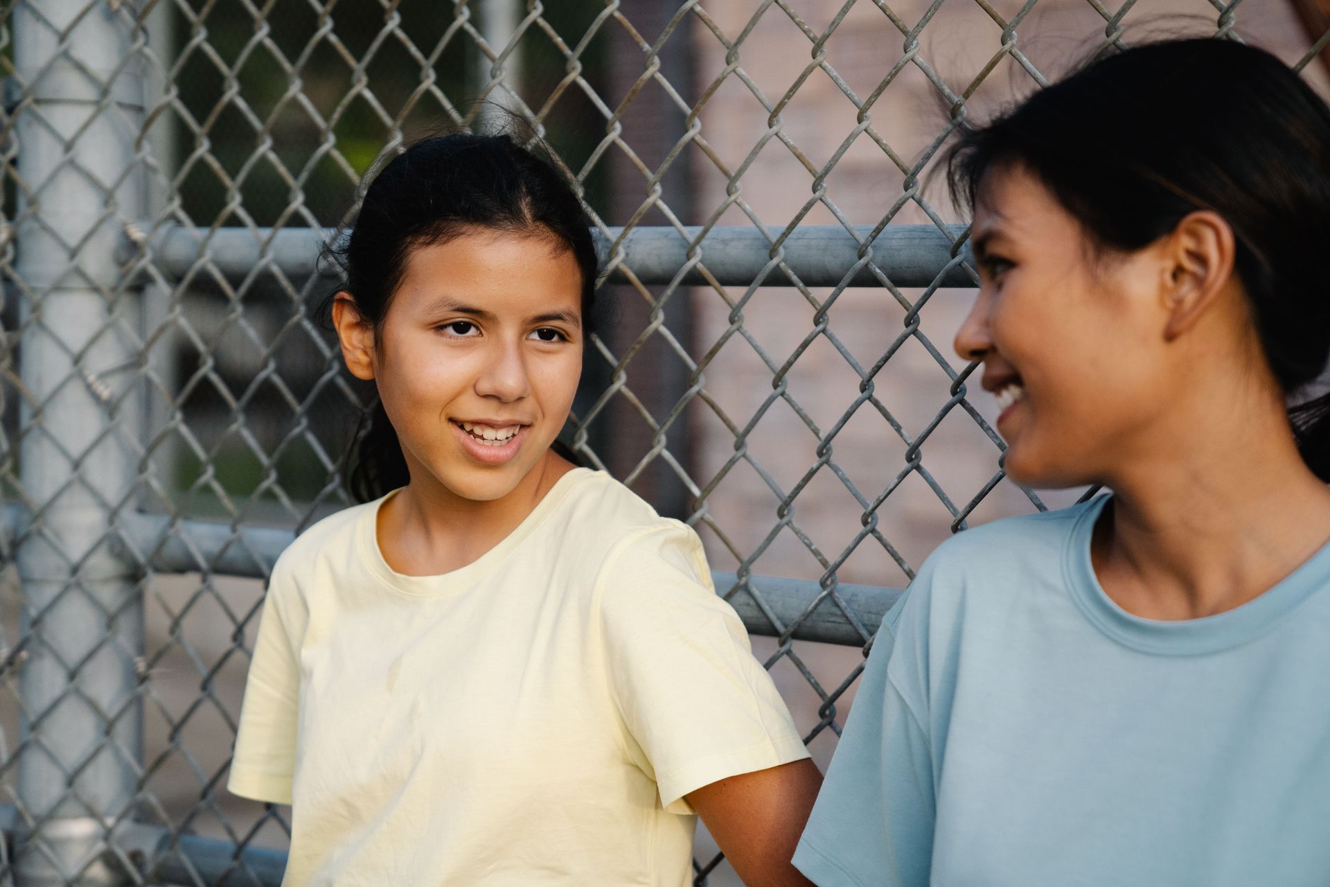 Two people smiling and talking in front of a chain-link fence
