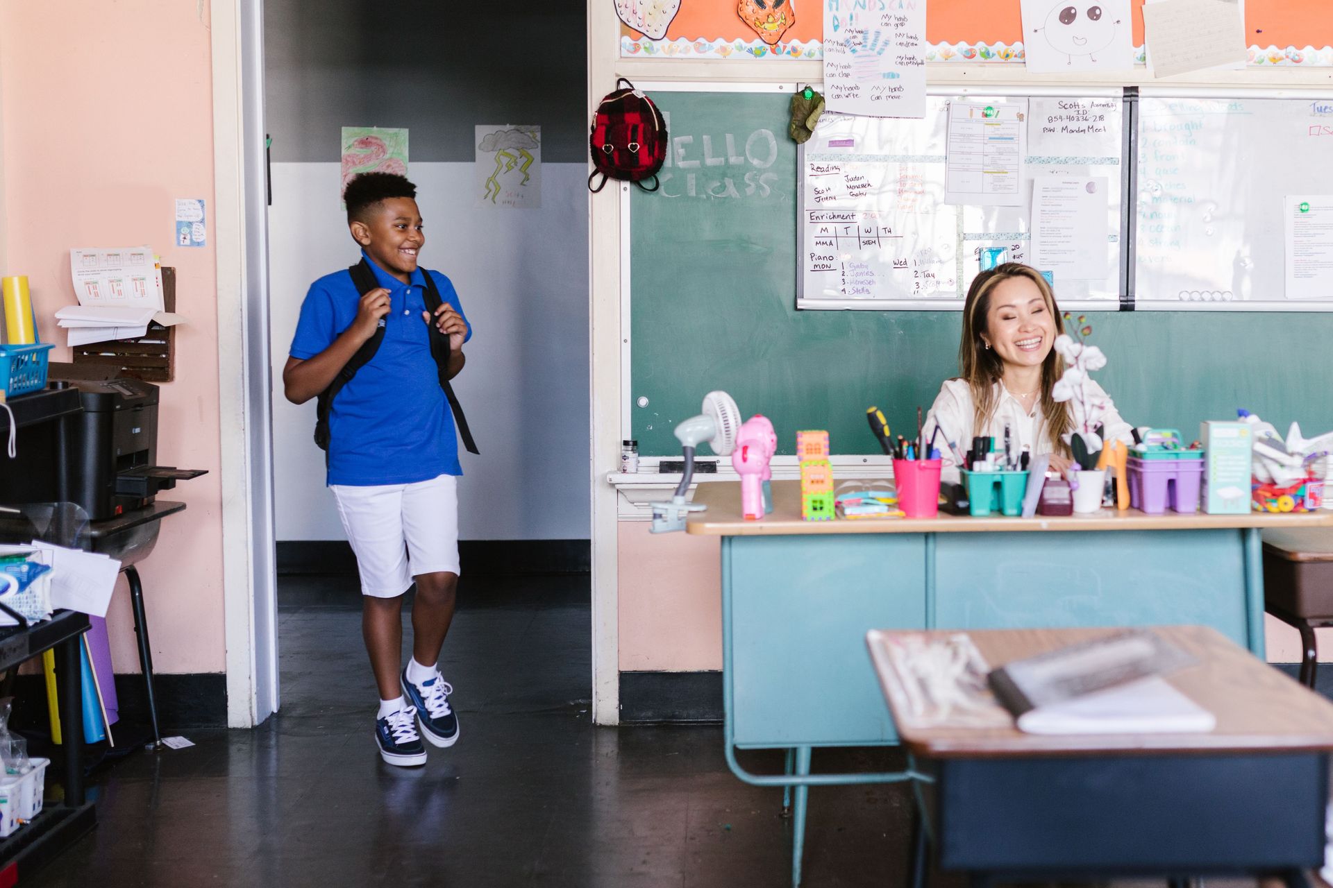 Classroom with a boy holding a backpack and a smiling  at a teacher seated at a desk