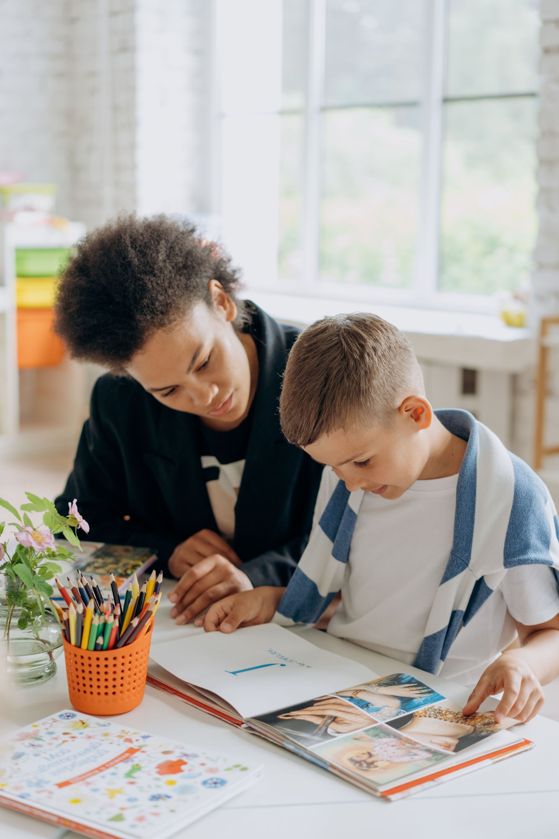 Adult and child reading together at a table with books, colored pencils, and drawings in a bright room