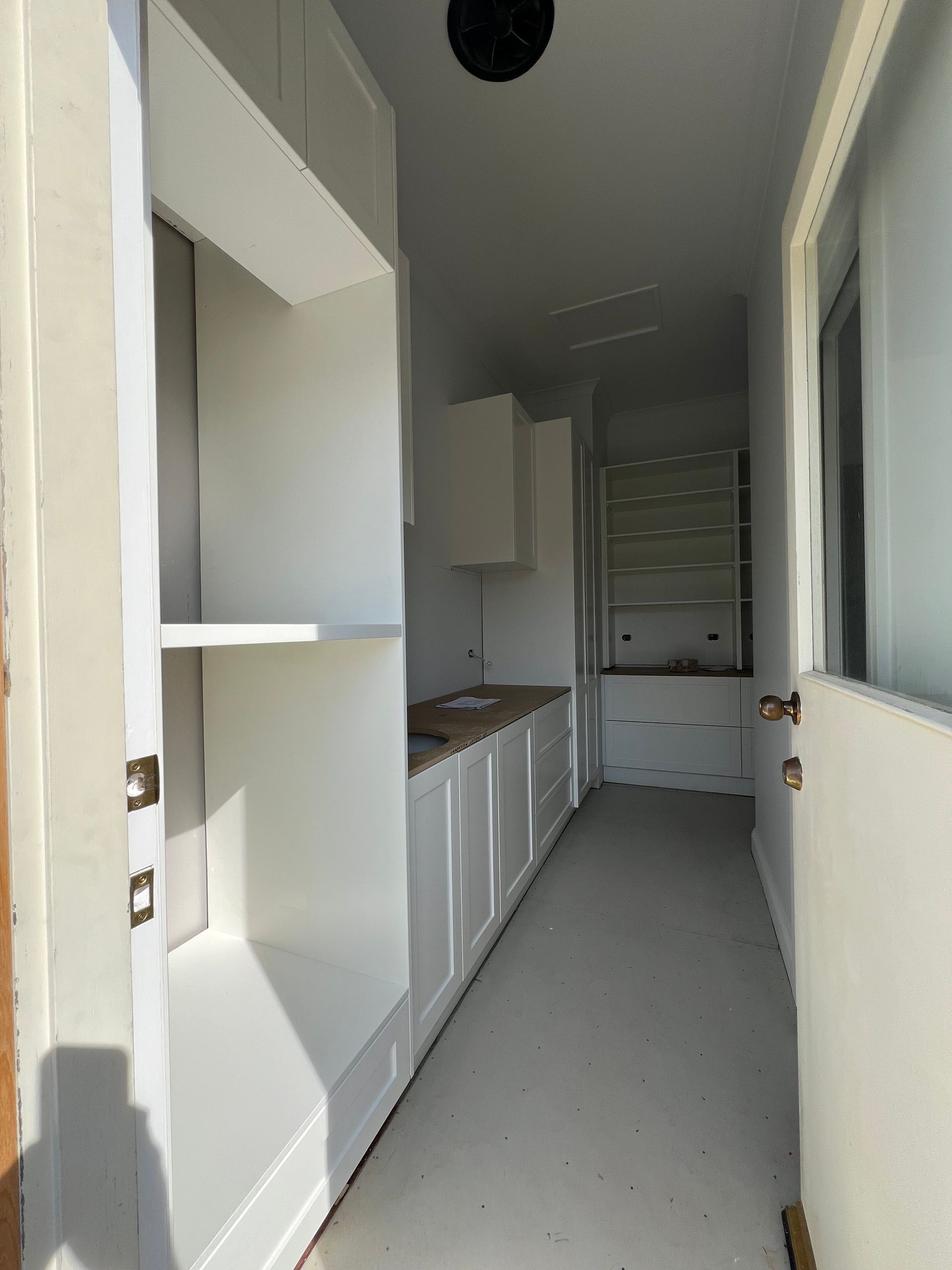 Narrow white galley kitchen with cabinets, countertop, window, and shelving.