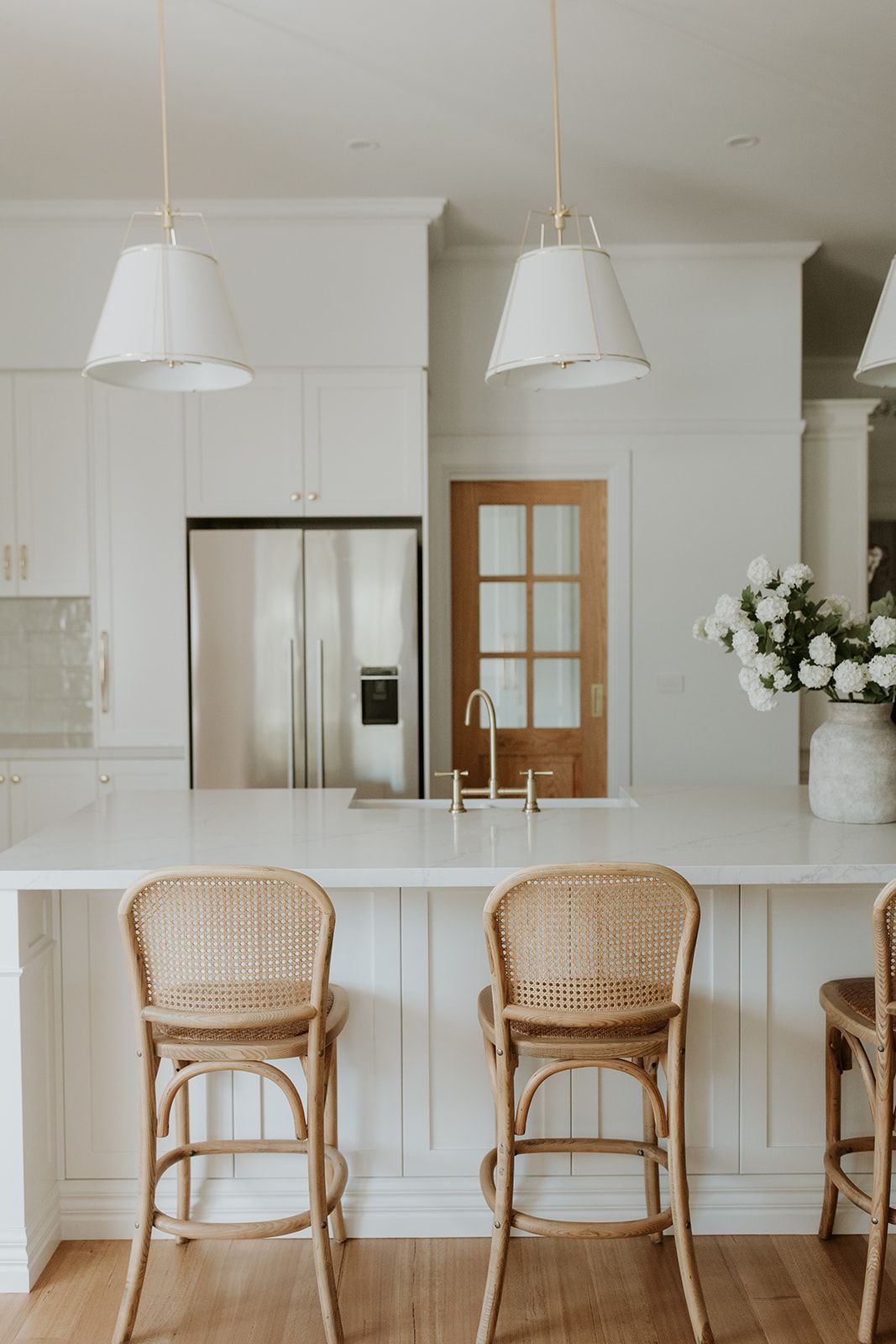Bright white kitchen with a marble island, wicker stools, pendant lights, and a stainless steel refrigerator.