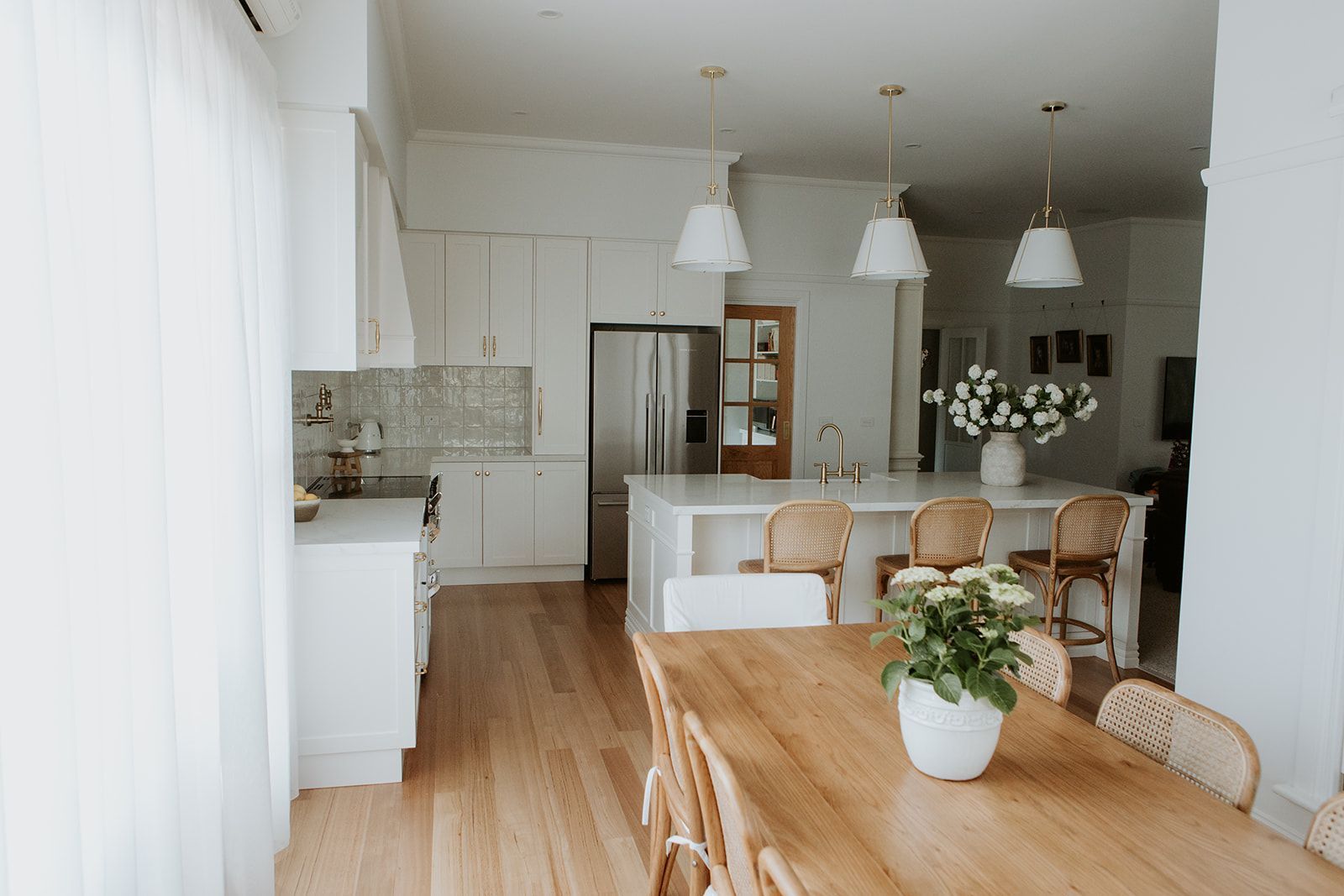 View from dining space to a modern country kitchen with rectangular island featuring stone benchtop and breakfast bar, with u-shaped cooking zone (left of fridge) and timber door to butler's pantry and laundry combination (right of  fridge).