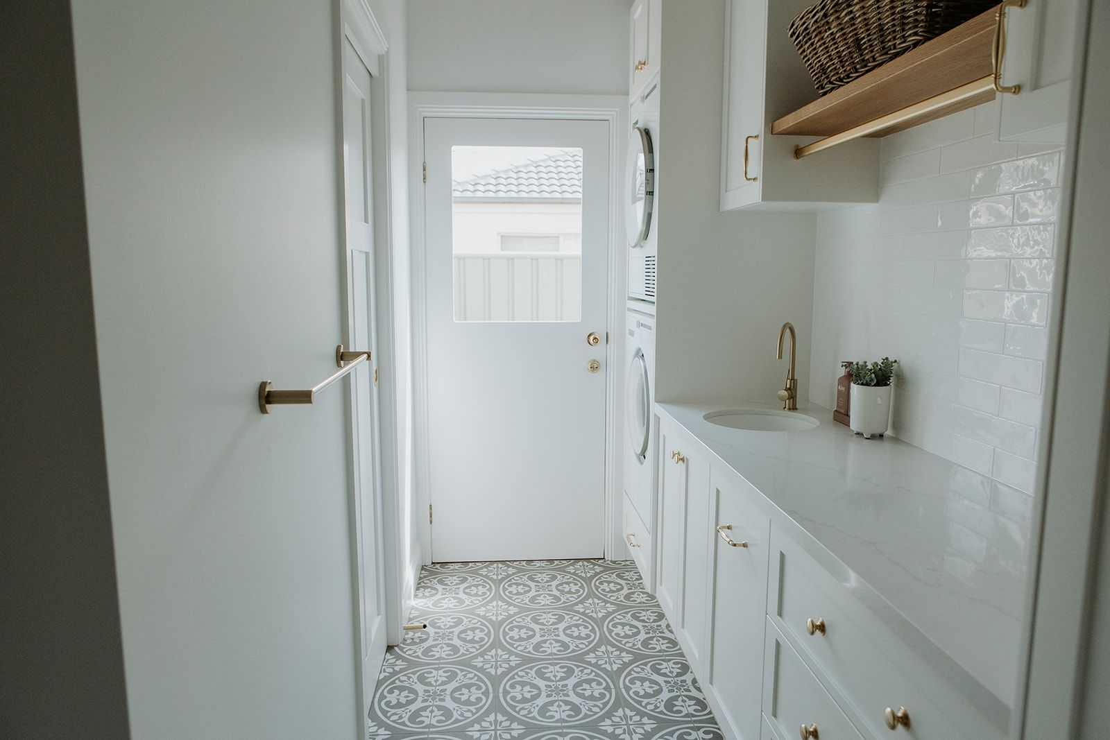 Narrow white hallway with built-in cabinets, brass fixtures, and patterned tile floor leading to a bright doorway