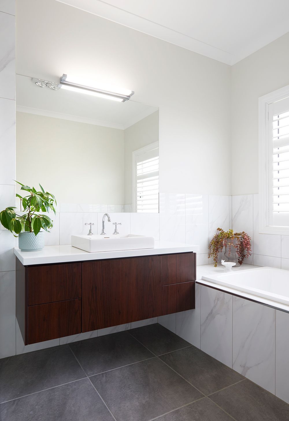 Modern bathroom with dark vanity, white countertop, bathtub, window shutters, and gray tile flooring