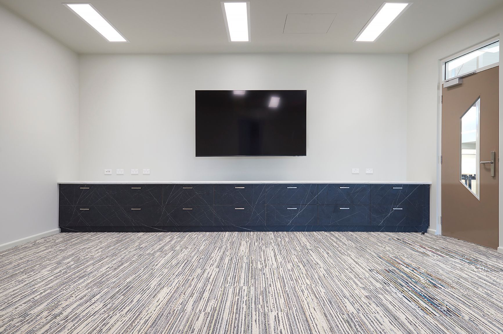 Empty modern room with wall-mounted TV, dark cabinetry, patterned carpet, and a glass door.
