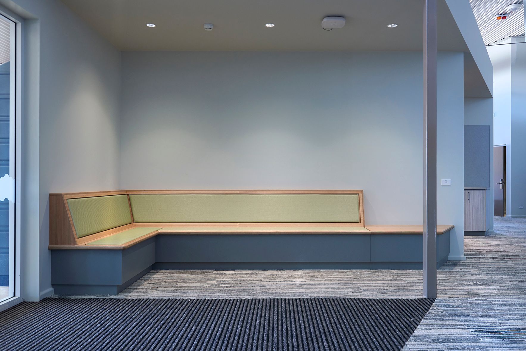 Empty modern office alcove with a built-in bench, green cushions, and gray carpet flooring