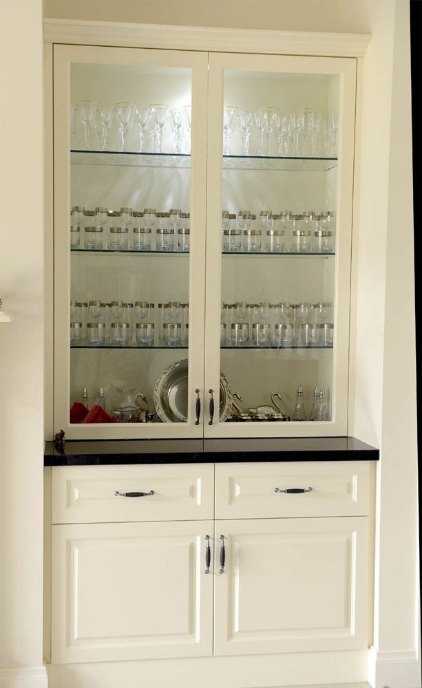 White built-in cabinet with glass doors, shelves of glassware, and lower drawers with silver handles