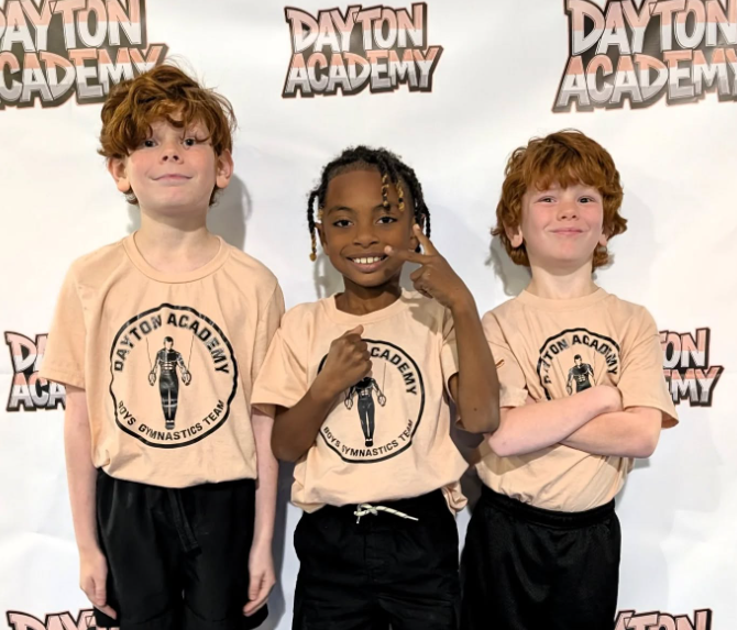 Three children posing in beige Dayton Academy shirts in front of a Dayton Academy backdrop