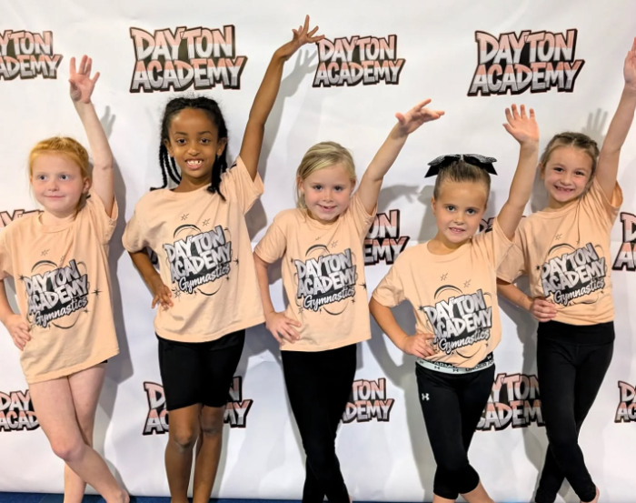 Five children in tan shirts pose with raised arms in front of a Dayton Academy backdrop.