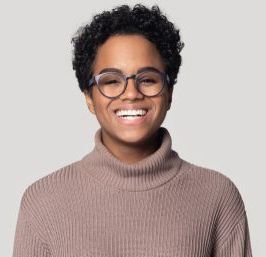 Smiling person with short curly hair and round glasses wearing a taupe turtleneck against a light gray background