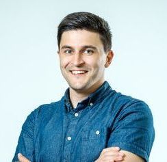 Smiling person in a blue button-up shirt with arms crossed against a light background