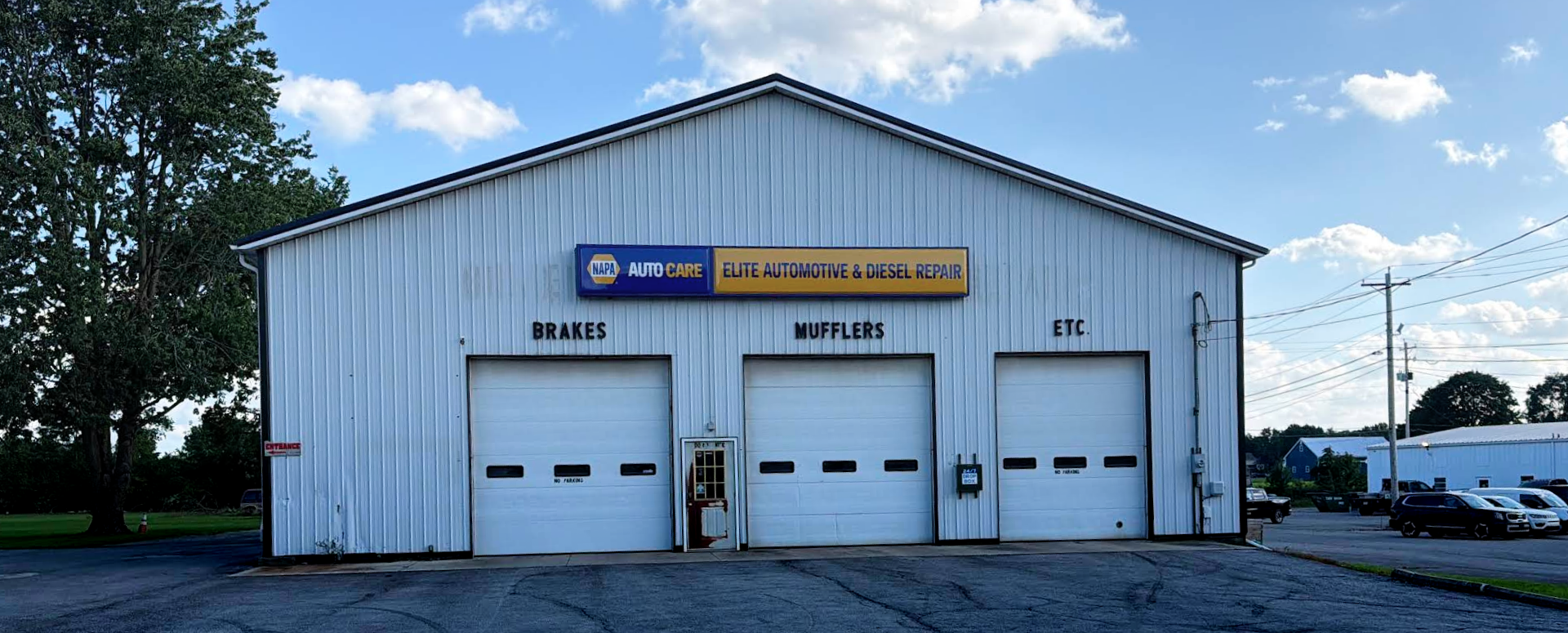 Front view of a white auto service building with three garage doors and a blue sign | Elite Automotive & Diesel Repair