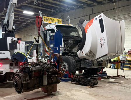 Disassembled truck and heavy equipment inside a repair garage, with a white cab lifted on a hoist.