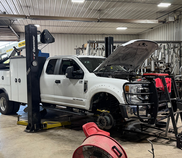 White pickup truck on a lift in an auto repair shop with hood open, surrounded by equipment and tools