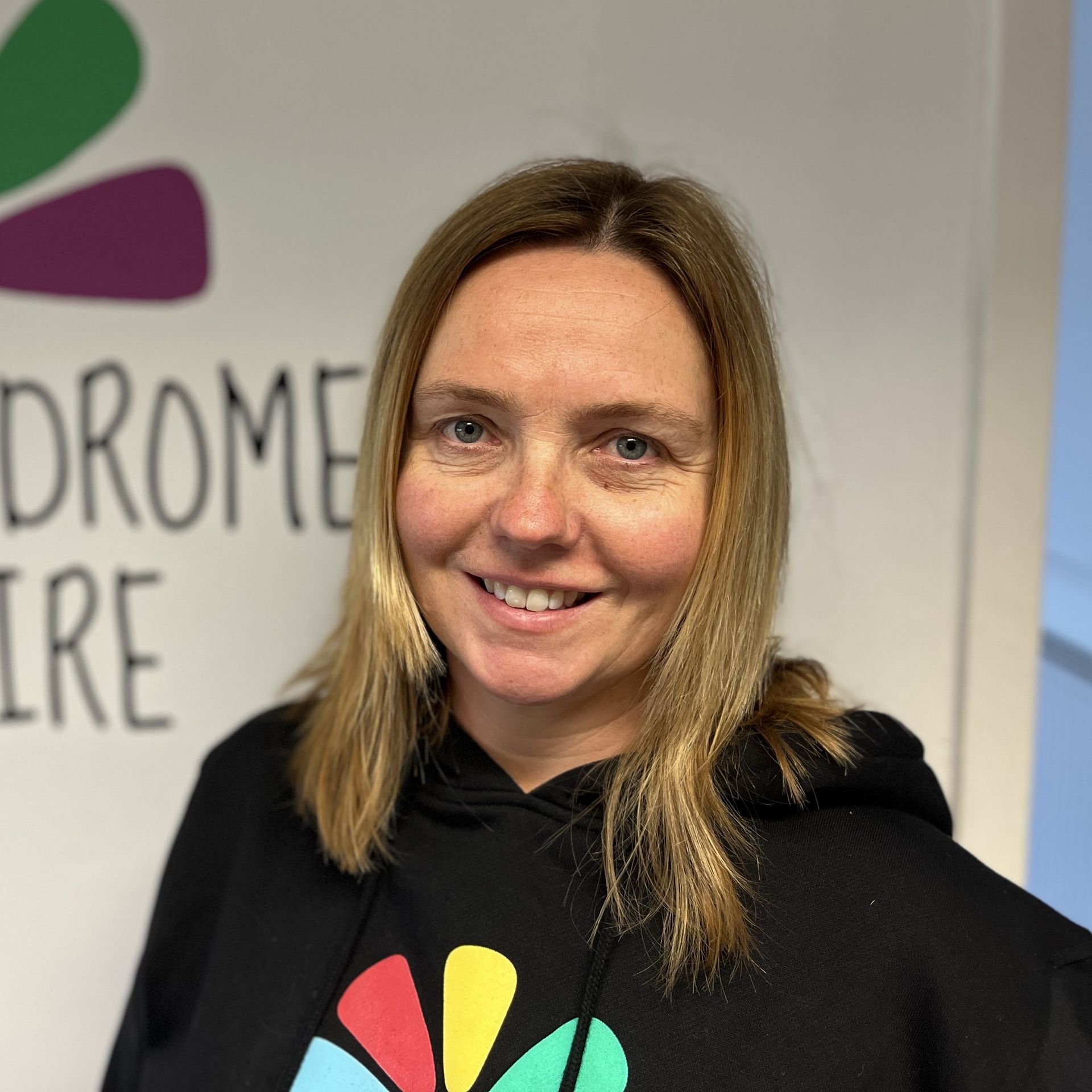 A woman in a black hoodie is smiling in front of a sign that says Down Syndrome Cheshire