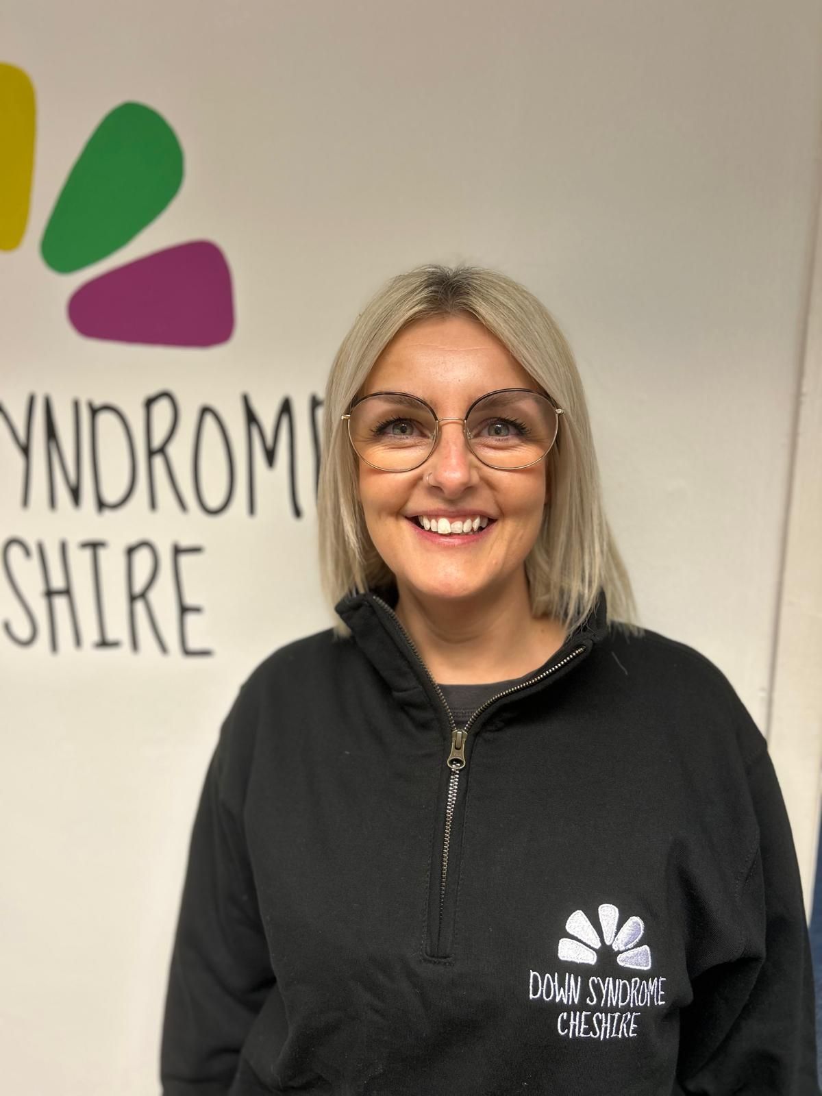 a woman wearing glasses is smiling in front of a sign that says Down Syndrome Cheshire