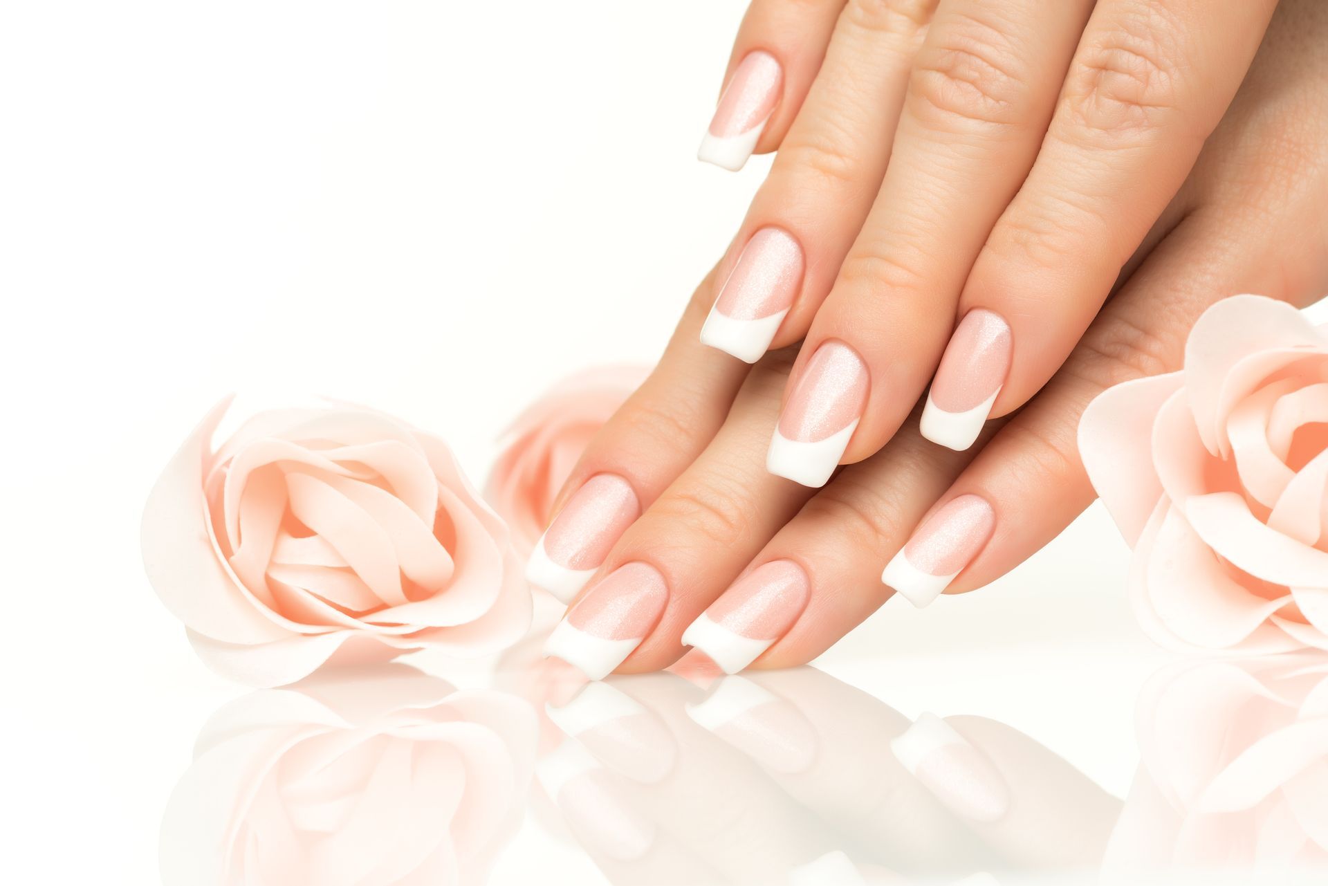 Manicured hands with white nails beside pale pink roses on a bright reflective surface