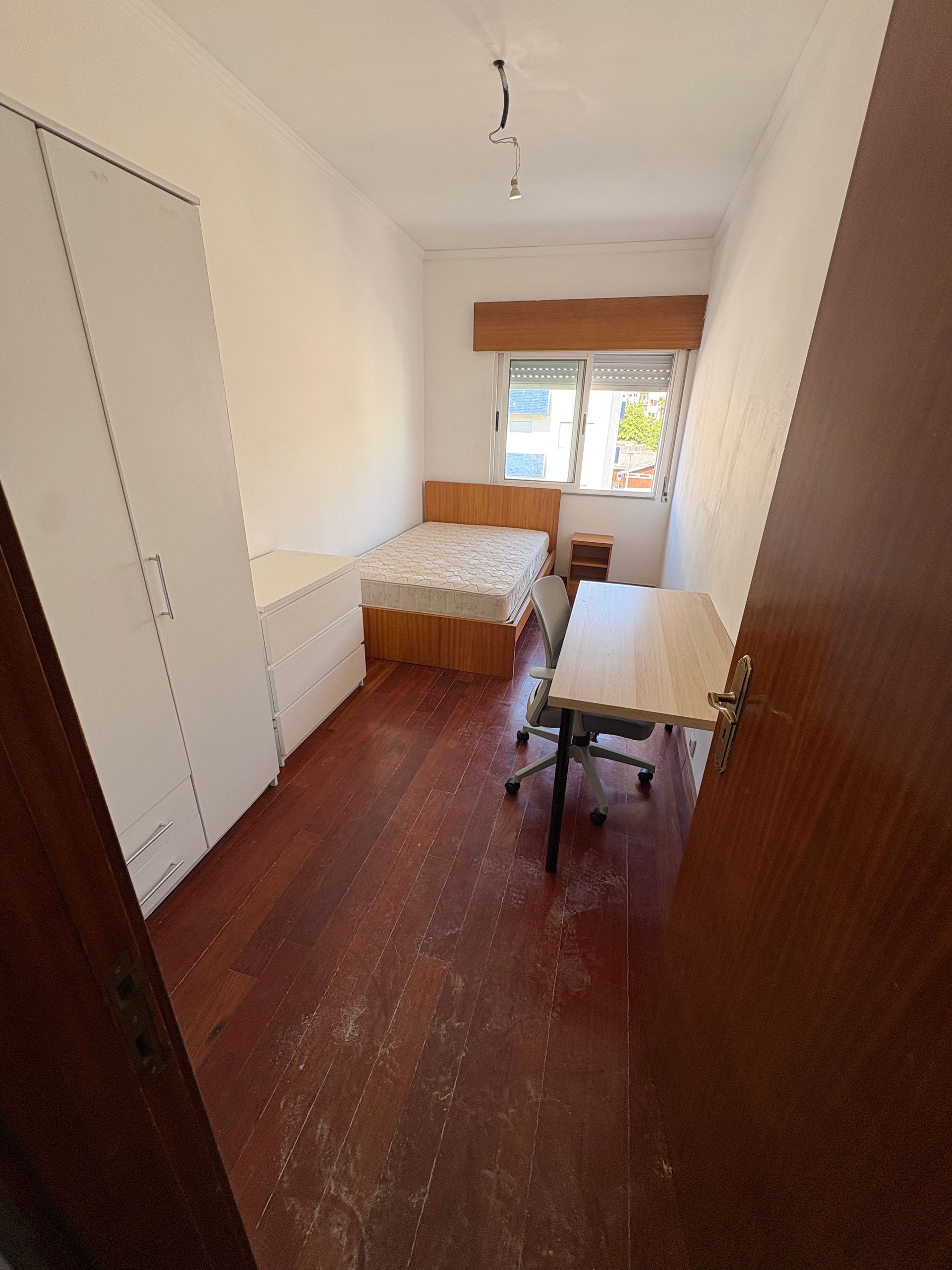 Narrow white bedroom with a single bed, table, chair, and window at the far end.