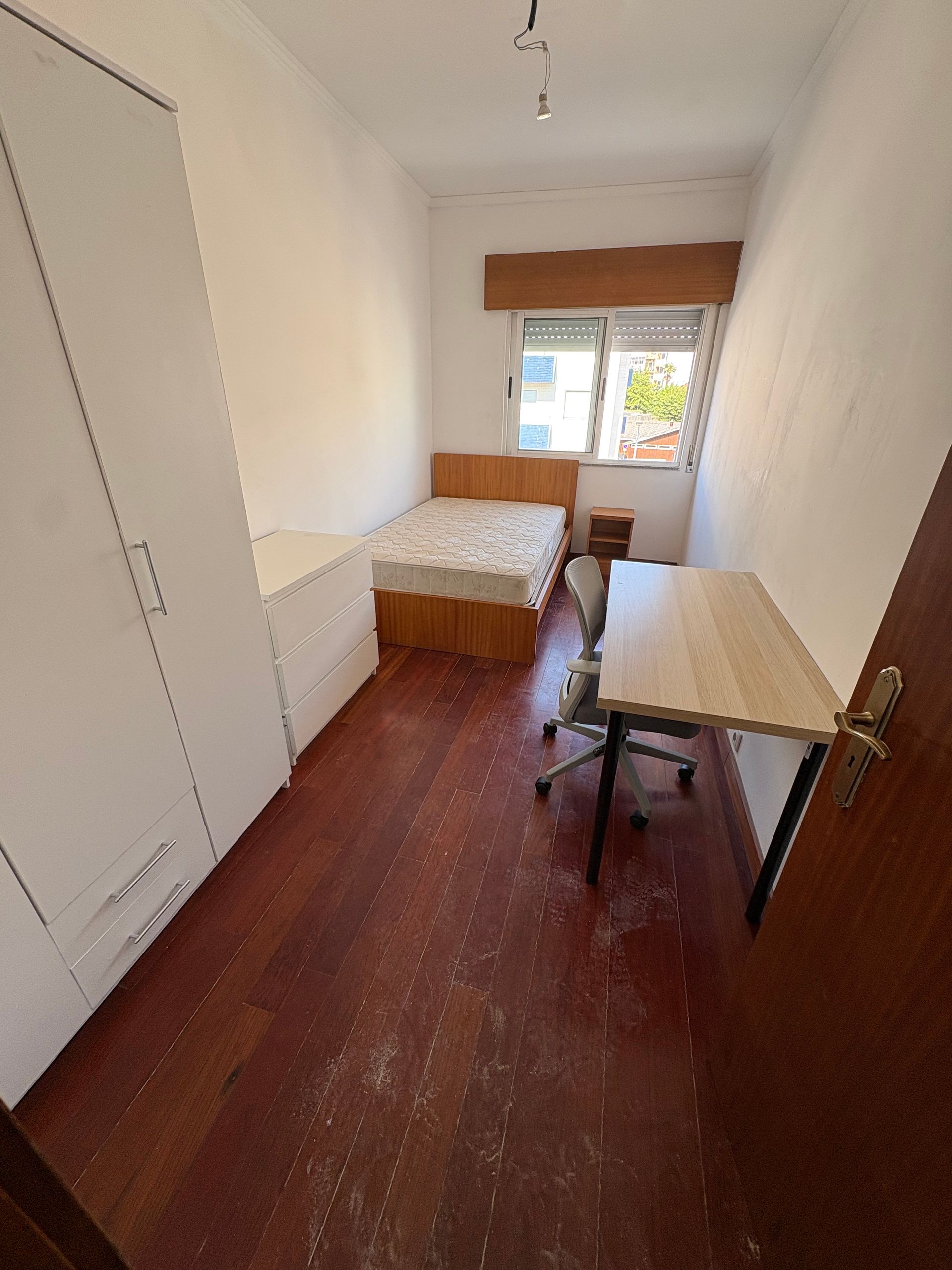 Narrow bedroom with single bed, desk, chair, and window at the far end.