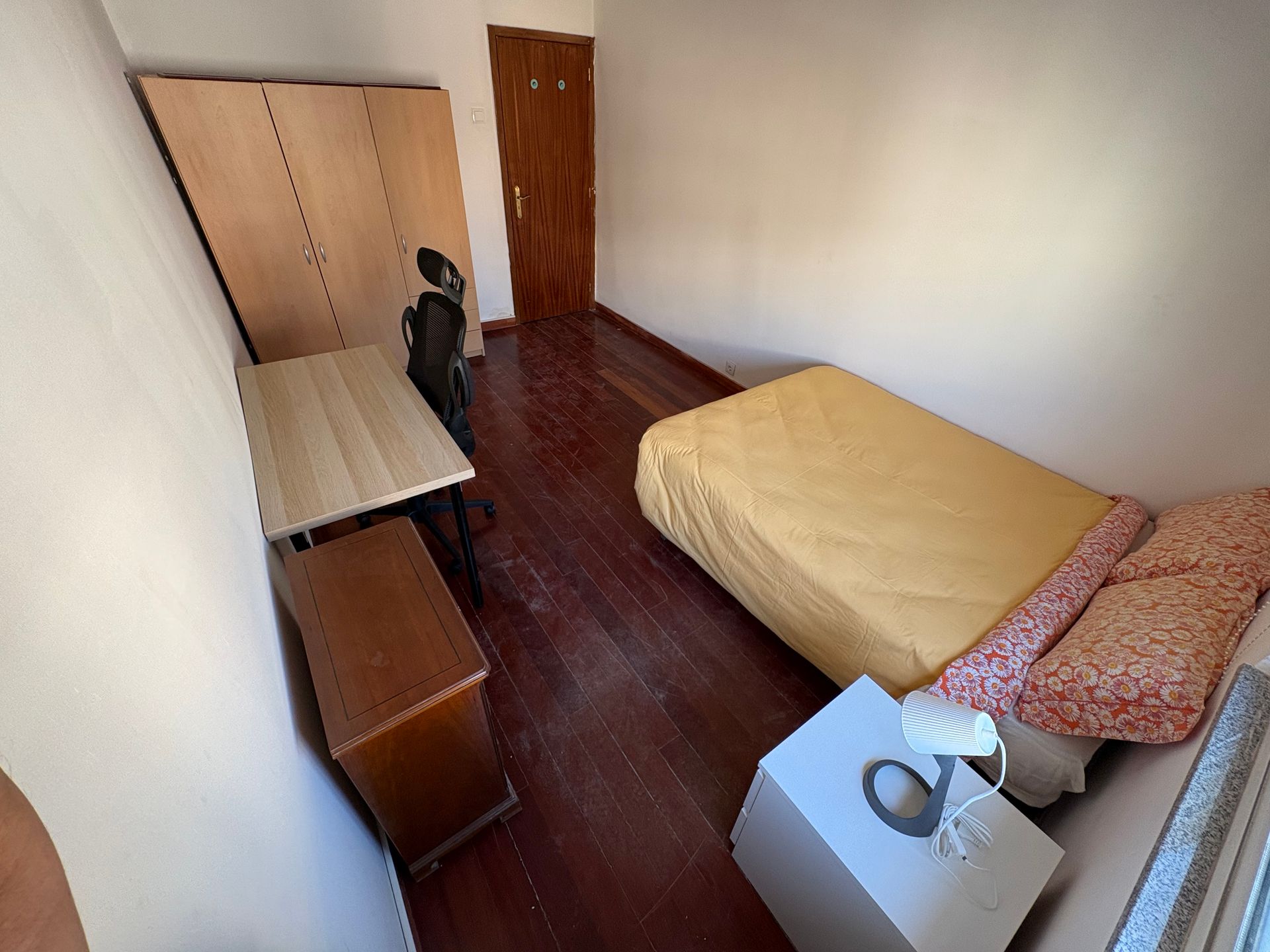 Small bedroom with bed, desk, wardrobe, chair, and a window; a cat sits near the wardrobe.