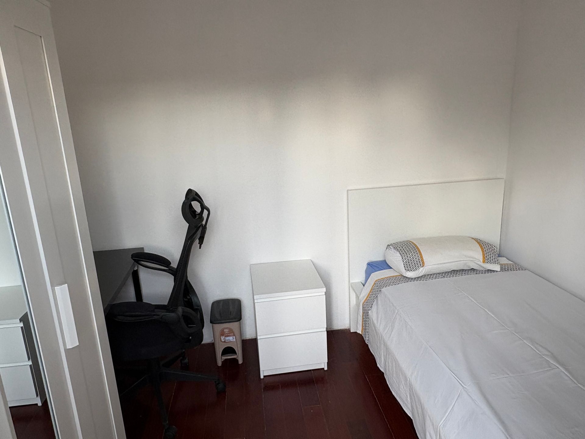 Small bedroom with a bed, nightstand, office chair, and trash can by a window.