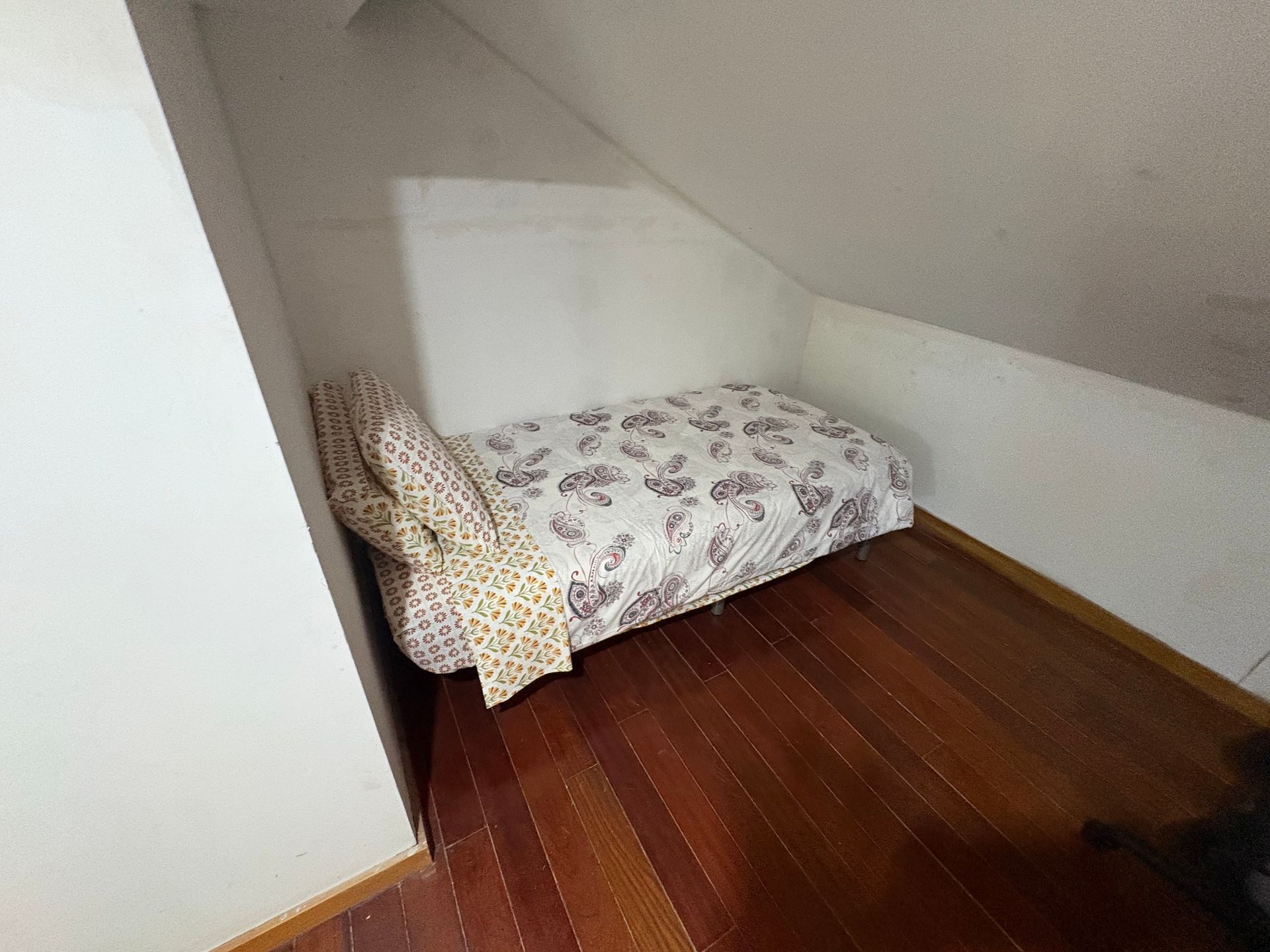 Small bed with floral bedding in a sloped-ceiling attic room with wooden floor.