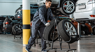 Mechanic in blue coveralls pushing a cart with two tires inside a garage, smiling. Cars in background. | Allen's Automotive Service