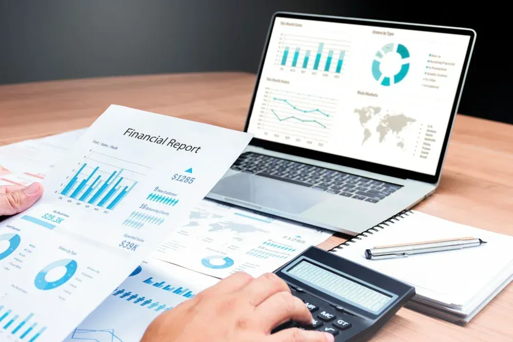 A person analyzes financial reports and data visualizations on a laptop screen with a calculator on a wooden desk.