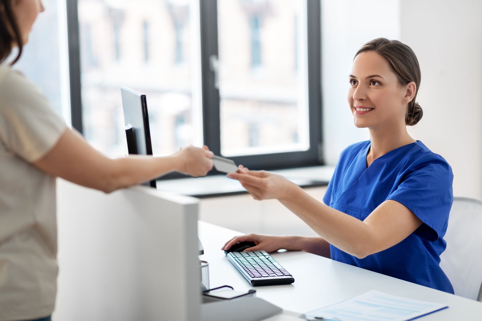 Woman in blue scrubs smiles, receiving card from patient at a reception desk. Woman in blue scrubs smiles, receiving card from patient at a reception desk.